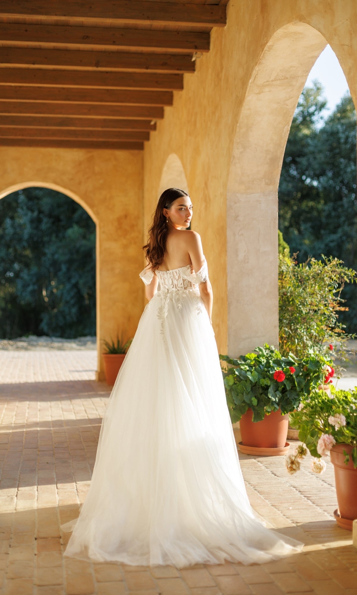 A woman in the Tina Holly TW480, a long formal off-shoulder white gown with a corset bodice, stands under a sunlit archway and looks over her shoulder, surrounded by potted plants and greenery.
